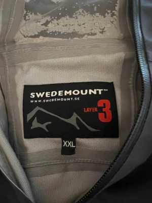 Grå vindjacka från Swedemount XXL - Säljer en grå vindjacka från Swedemount i storlek XXL. Jackan har dragkedja, detaljer i limegrönt och Swedemount-logga på bröstet. Materialet är lätt och vindtätt, perfekt för utomhusaktiviteter. Snygg och funktionell design med sportig känsla.