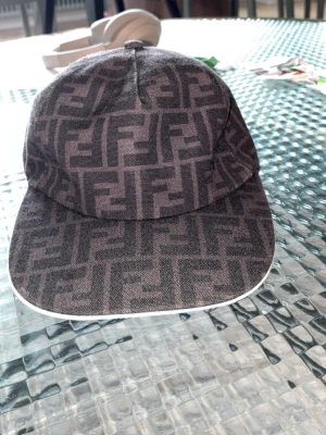 Fendi keps med FF-mönster - Snygg keps från Fendi med det klassiska FF-logomönstret i svart och grått. Kepsen har böjd skärm med vit kant och är tillverkad i bomullsmix. Perfekt accessoar för att lyfta din streetwear-look.