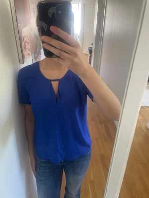 Blå topp med cut out från Stockh lm - Snygg blå topp från Stockh lm med korta ärmar och unik cut out-detalj vid halsen. Tillverkad i ett mjukt, glansigt material som ger en stilren look. Perfekt att matcha med jeans eller kjol för en trendig outfit.