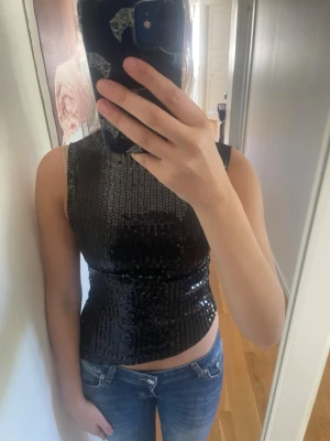 Svart paljett-topp med hög hals - Säljer en svart croppad topp med paljetter och hög hals. Toppen är ärmlös och har en slim passform som framhäver figuren. Perfekt för dig som vill sticka ut med lite extra glitter. Materialet är polyester och toppen har en snygg glansig finish.