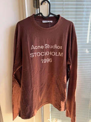 Choklad Brun långärmad tröja Acne Studios - Cool choklad brun långärmad tröja från Acne Studios med vit text 'Acne Studios STOCKHOLM 1996' på bröstet. Tröjan har rund halsringning och är gjord i mjukt bomullsmaterial. Perfekt för en avslappnad och trendig look.