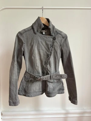 Grå jeansjacka med bälte och asymmetrisk knäppning - Snygg grå jeansjacka med unik asymmetrisk knäppning och tillhörande bälte i midjan. Jackan har hög krage, långa ärmar och markerad midja för en cool och modern siluett. Perfekt för dig som vill sticka ut med en annorlunda jeansjacka. paketpris om du köper fler