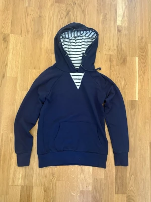 Mörkblå hoodie med randig huva - Snygg mörkblå hoodie med känguruficka och huva som har ett vitt och marinblått randigt foder. Klassisk passform med ribbade muddar vid ärmslut och nederkant. Perfekt för en chill och avslappnad stil.