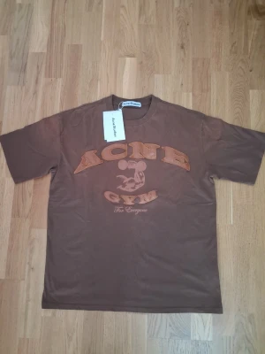 Brun Acne Studios t-shirt med tryck - Cool brun t-shirt från Acne Studios med trycket 'ACNE GYM' och illustration framtill. Klassisk passform med korta ärmar och rund hals. Perfekt för en avslappnad och trendig stil.