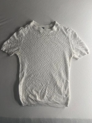 Vit spetsstickad topp från H&M - Vit kortärmad topp från H&M med spetsstickat diamantmönster och ribbad krage. Toppen är i bomull och har en feminin, luftig känsla som passar perfekt till jeans eller kjol. Snyggt mönster och klassisk passform.