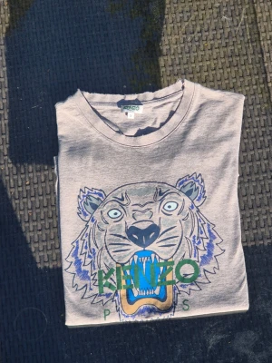 Grå Kenzo t-shirt med tigertrycket - Säljer en grå t-shirt från Kenzo med det ikoniska tigertrycket i blått, grönt och beige på framsidan. . Perfekt för dig som vill sticka ut med en cool och trendig look. Den är i bra skick för att vara köpt på nk 2023 nypris cirka 1000kr värderad till 400-700kr i detta skick varit inne på kemtvätt innan försäljning 