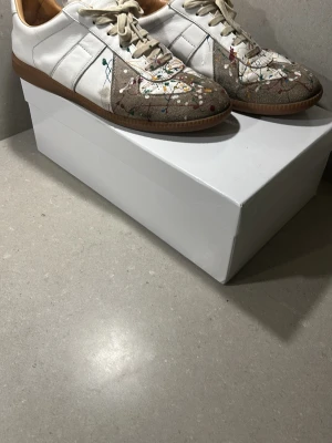 Maison Margiela sneakers med färgstänk - Unika sneakers från Maison Margiela i vitt och beige med färgstänk i flera färger. Skorna har klassisk låg siluett, vita snören och gummisula. Materialmix av läder och mocka ger en cool kontrast och streetkänsla. Perfekta för dig som vill sticka ut med din stil.