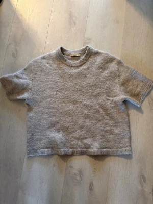 Ljusgrå stickad t-shirt från Arket - Mysig ljusgrå stickad t-shirt från gina med korta ärmar och rund halsringning. Tröjan har en avslappnad passform och ribbstickade detaljer vid ärmslut, hals och nederkant. 