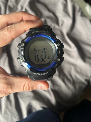 Svart digital sportklocka - Digital sportklocka från Football Watch med svart och blå design. Klockan har en rund display, flera knappar för olika funktioner och vattentålig konstruktion. Armbandet är i svart syntetmaterial och spännet är i metall. Perfekt för dig som gillar sportig stil.
