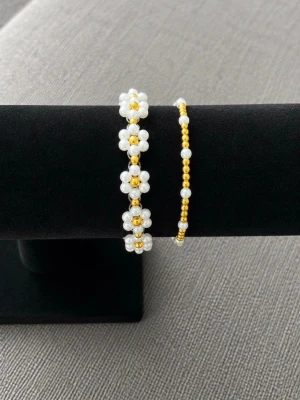 Två pärlarmband med gulddetaljer och en med blommor  -  två armband med vita och guldiga pärlor. Det ena har små blommor av pärlor och guldfärgade detaljer, det andra är enkelt med vita och guldiga pärlor i rad. 