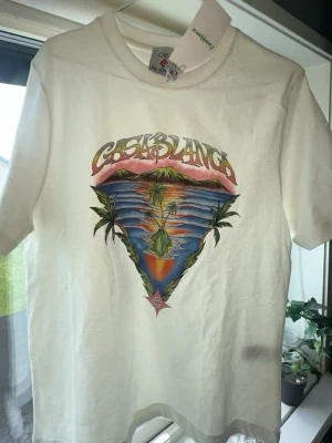 Vit t-shirt med tryck från Casablanca - Vit t-shirt från Casablanca med färgstarkt tryck av solnedgång, palmer och hav på bröstet. T-shirten har rund hals och korta ärmar. Materialet är mjuk bomull och passformen är normal. Perfekt för dig som gillar unika prints och coola märken.