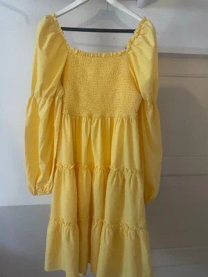 Gul volangklänning med puffärmar - Söt gul klänning med volanger och långa puffärmar. Klänningen har smock upptill och en luftig, flowy passform. Perfekt för soliga dagar och ger en riktigt somrig vibe.