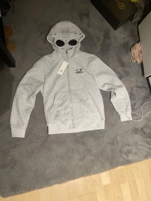 Grå hoodie från C.P. Company - Snygg grå hoodie från C.P. Company med dragkedja och ikoniska glasögon i huvan. Broderad logga på bröstet och två fickor framtill. Perfekt för dig som gillar streetwear och vill sticka ut lite extra.
