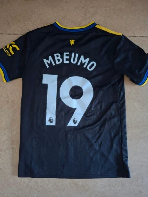 Manchester United bortatröja svart/gul - Snygg Manchester United bortatröja från Adidas i svart med gula detaljer och blå inslag. Tröjan har trycket 'MBEUMO 19' på ryggen, klubbmärke och sponsorlogga på bröstet samt gula ränder på axlarna. Tillverkad i lätt och ventilerande material.