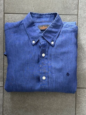 Blå linneskjorta från Morris - Snygg blå skjorta från Morris i linne med klassisk krage och vita knappar. Skjortan har lång ärm och en liten broderad logga på bröstet. Perfekt för en stilren och avslappnad look.