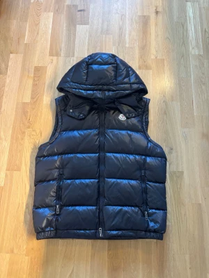 Svart dunväst med huva från Moncler - Snygg svart dunväst från Moncler med avtagbar huva och två dragkedjefickor framtill. Västen har quiltad design och Moncler-logga på bröstet. Perfekt för lager-på-lager och håller dig varm med stil. Väldigt sugen på byten! Pris kan diskuteras. Säljs billigt då jag är osäker på äkthet!!