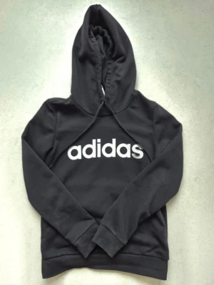 Svart hoodie från Adidas - Svart hoodie från Adidas med stor vit logga framtill. Klassisk modell med huva och dragsnören, samt långa ärmar och ribbade muddar. Perfekt för en avslappnad och sportig stil. Tillverkad i mjukt material som känns skönt mot huden.