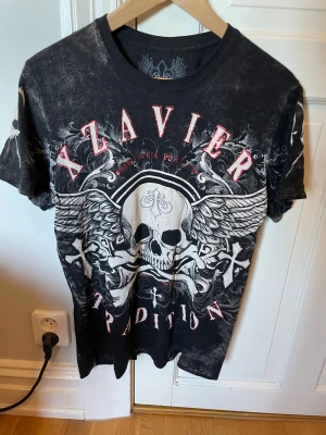 Svart Xzavier t-shirt  - Cool svart t-shirt från Xzavier med stort dödskalletryck och vingar både fram och bak. T-shirten har korta ärmar och detaljerade grafiska mönster i vitt och rött. Lowk edgy?