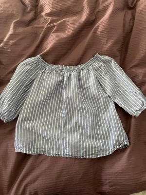 Randig offshoulder blus i blått - Snygg blåvit randig blus med offshoulder-modell och trekvartsärm. Blusen har resår vid axlarna och är tillverkad i ett lätt och luftigt tyg.