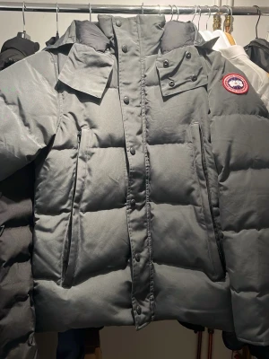 Grå dunjacka från Canada Goose - Snygg grå dunjacka från Canada Goose med klassisk patch på ärmen. Jackan har hög krage, avtagbar huva och stora fickor framtill. Perfekt för kalla vinterdagar och riktigt varm tack vare dunfyllningen. Stängs med dragkedja och knappar. Priset inte hugget i sten.