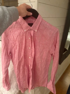 Rosa linneskjorta från Oscar Jacobson - Snygg rosa skjorta från Oscar Jacobson i linnematerial. Skjortan har klassisk krage med knappar, vita knappar framtill och lång ärm. Passformen är slim och skjortan har en stilren, enkel design som passar till många tillfällen.
