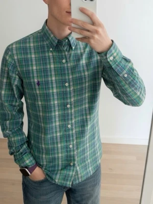 Grön rutig skjorta från Ralph Lauren - Snygg grön och blå rutig skjorta från Ralph Lauren med klassisk button-down krage och lila broderad logga på bröstet. Skjortan har långa ärmar och vita knappar framtill. Perfekt för en stilren och avslappnad look.