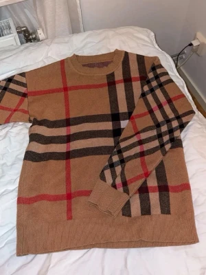 Burberry tröja  - Stickad tröja i beige med klassiskt rutigt mönster i svart, rött och brunt. Tröjan har rund halsringning och ribbade muddar vid ärmslut och nederkant. Perfekt för dig som gillar stilrena och trendiga plagg.