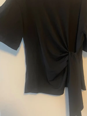 Svart topp med knyt i midjan - Trendig svart topp med korta ärmar och snygg knytning i midjan. Materialet är stretchigt och svalt, nästan som träningsmaterial. Det sitter super snyggt på. Endast andvänd en gång så den är i mycket bra skick. Säljer då jag inte får någon andvändning för den.