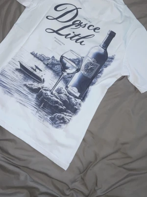 Vit t-shirt med vintagemotiv - Snygg vit t-shirt med stort tryck på ryggen som visar en vinflaska, ett vinglas och en båt vid kusten. Texten 'Doce Lita' syns i elegant stil. Perfekt för dig som gillar unika motiv och chill vibes.