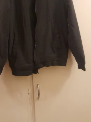 Svart bomberjacka från Lyle & Scott - Stilren svart bomberjacka från Lyle & Scott med dragkedja framtill och två snedställda fickor. Klassisk passform och ribbade muddar vid ärmslut och midja. Passar till vår/höst eller mild vinter. Mörkt grå, nästan svart.