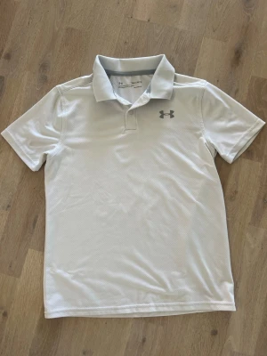 Vit pikétröja från Under Armour, stl junior M - Snygg vit pikétröja från Under Armour med klassisk krage och korta ärmar. Tröjan har en diskret logga på bröstet och är tillverkad i ett lätt och svalt syntetmaterial som andas. Perfekt för en sportig och stilren look på golfbanan⛳️🏌🏼‍♂️! 