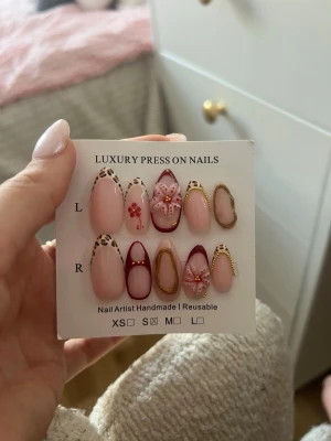 Lyxiga press-on naglar med leopard och blommor - Säljer ett set lyxiga press-on naglar i mandelform med nude bas, leoparddetaljer, guldglitter och handmålade blommor. Mix av rosa, nude och vinröda toner. Naglarna är handgjorda och återanvändbara, perfekta för dig som vill ha unika och trendiga naglar. Det ingår ett kitt med nagellim, nagelklister, nagelfil, nagelbands pusher
