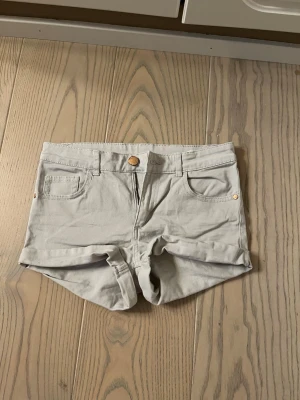 Ljusbeige höga shorts med fickor - Snygga ljusbeige shorts med hög midja och klassisk femficksdesign. De har uppvikta benslut, bälteshällor och stängs med knapp och dragkedja. Perfekta för varma dagar och enkla att matcha med olika toppar.