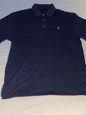 Mörkblå pikétröja från Ralph Lauren - Klassisk mörkblå pikétröja från Ralph Lauren med vit broderad logga på bröstet. Tröjan har krage, tre knappar framtill och är tillverkad i mjuk bomull. Perfekt till en avslappnad men ändå stilren look.