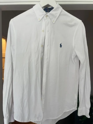 Vit skjorta från Ralph Lauren - Klassisk vit skjorta från Ralph Lauren med blå broderad logga på bröstet. Skjortan har lång ärm, knappar framtill och button-down krage. Perfekt för en clean och stilren look.