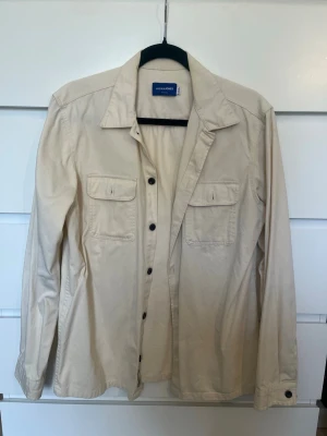 Beige overshirt från Jack & Jones - Stilren beige overshirt från Jack & Jones med klassisk krage, svarta knappar och två bröstfickor med lock. har långa ärmar med knapp vid ärmslut. Perfekt för lager på lager och enkel att matcha.