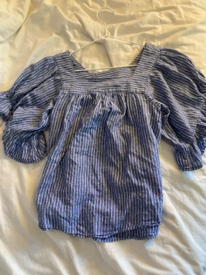 Blårandig blus med puffärmar - Snygg blårandig blus med bred fyrkantig ringning och puffiga korta ärmar. Blusen har resår vid ärmslut och nederkant för en trendig look. Perfekt att styla med jeans eller kjol för en cool vardagsstil.