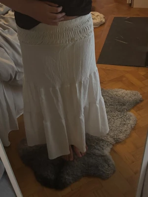 Vit långkjol med smock i midjan - Säljer en vit långkjol med bred smock i midjan och volangdetaljer. Kjolen har en luftig och flowy känsla, perfekt för varmare dagar. Enkel att matcha med olika toppar för en chill och stilren look.