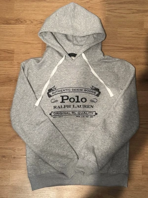 Grå hoodie från Polo Ralph Lauren - Snygg grå hoodie från Polo Ralph Lauren med svart tryck framtill och vita dragsnören i huvan. Klassisk passform med ribbade muddar och mjukt material som känns skönt mot huden. Perfekt för en avslappnad och trendig stil.