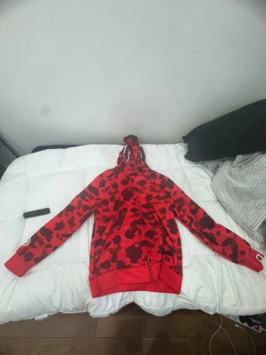 Röd camo hoodie från BAPE - Säljer en röd hoodie från BAPE med ikoniskt camo-mönster i svart och mörkrött. Hoodien har dragkedja hela vägen, huva och en vit stjärnlogga på ärmen. Perfekt för dig som vill sticka ut med streetwear-stil.