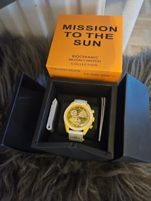 Omega x Swatch Mission to the Sun - OBS. Boxen är trasig! Snygg Omega x Swatch Mission to the Sun från Bioceramic MoonSwatch Collection. Klockan har gul boett, vit urtavla med gula och svarta detaljer samt vit rem med Omega-logga. Kommer i originalförpackning med tillbehör. Perfekt statement piece för dig som gillar unika accessoarer.