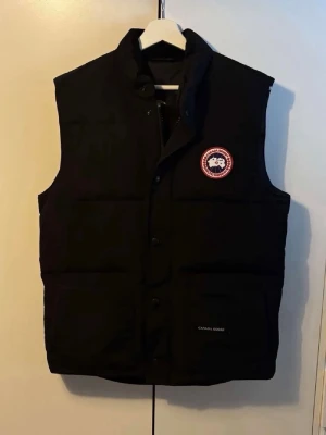 Svart dunväst från Canada Goose - Snygg svart dunväst från Canada Goose med klassisk logotyp på bröstet. Västen har hög krage, knäppning med tryckknappar och dragkedja samt två fickor framtill. Perfekt för lager-på-lager och håller dig varm under kyliga dagar.