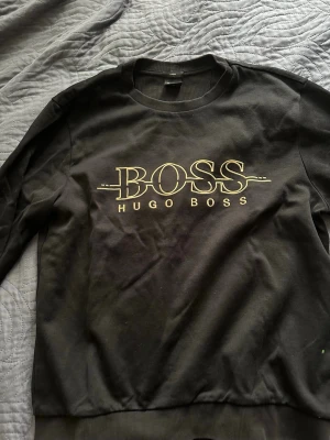 Svart sweatshirt från Hugo Boss - Snygg svart sweatshirt från Hugo Boss med stort guldfärgat BOSS-tryck på bröstet. Klassisk rund halsringning och ribbade muddar vid ärmslut och nederkant. Perfekt för en stilren och avslappnad look.