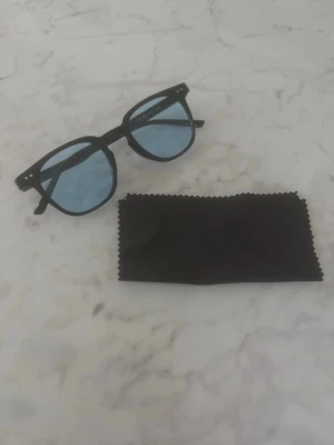 Svarta solglasögon med blåtonade glas - Snygga solglasögon med svart båge och runda blåtonade glas. Klassisk design som passar till de flesta outfits. Medföljer en svart putsduk för enkel rengöring. Perfekta för soliga dagar och ger en cool vibe. Helt nya och oanvända!
