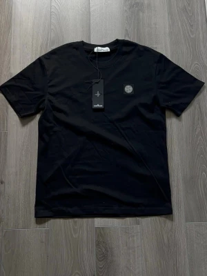 Svart t-shirt från Stone Island - Snygg svart t-shirt från Stone Island med klassisk rund hals och korta ärmar. T-shirten har en liten patch med logga på bröstet och är tillverkad i mjuk bomull för en skön känsla. Perfekt till jeans eller shorts.