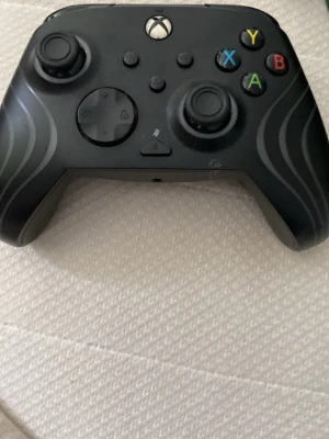 Svart Xbox handkontroll - Säljer en svart Xbox handkontroll med klassisk layout, två analoga spakar, styrkors och färgade knappar (A, B, X, Y). Handkontrollen har ergonomisk form och detaljerade linjer på framsidan. Perfekt för gaming på konsol eller PC.