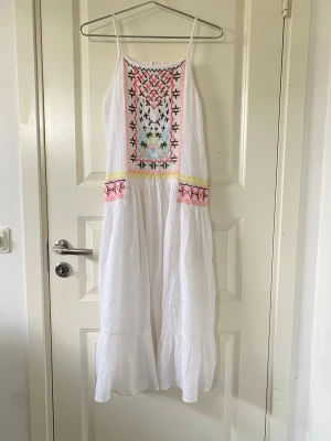 Vit långklänning med broderi - Luftig vit långklänning med tunna axelband och färgglada broderade detaljer framtill i rosa, svart, blått och gult. Klänningen har volang nedtill och är tillverkad i ett lätt bomullstyg. Perfekt för varma sommardagar.