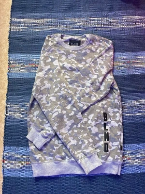 Grå camomönstrad hoodie från Blend - Snygg grå tröja från Blend med camomönster i olika grå nyanser och BLEND-tryck på sidan. Tröjan har ribbade muddar vid ärmslut och nederkant samt en klassisk avslappnad passform. Perfekt för en chill och street-inspirerad stil.