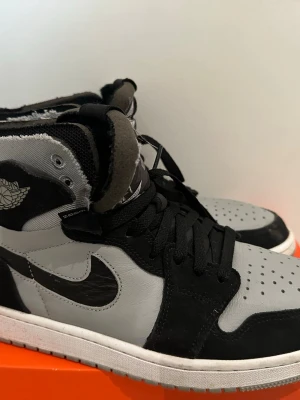 Nike Air Jordan 1 Zoom svart/grå - Säljer ett par Nike Air Jordan 1 Zoom sneakers i svart och grått med högt skaft. Skorna har klassisk Jordan-logga på sidan, Zoom Air-märke på tungan och perforerad tå. Materialet är en mix av läder och textil, med svarta skosnören och vit sula.