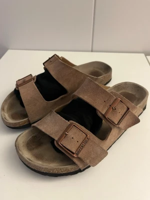 Beige mockasandaler från Birkenstock - Snygga sandaler från Birkenstock i beige mocka med två breda remmar och justerbara metallspännen. Klassisk fotbädd och svart sula för extra komfort. Perfekta för en avslappnad och trendig look.